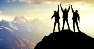Silhouette of three people triumphantly holding hands after reaching a mountain summit